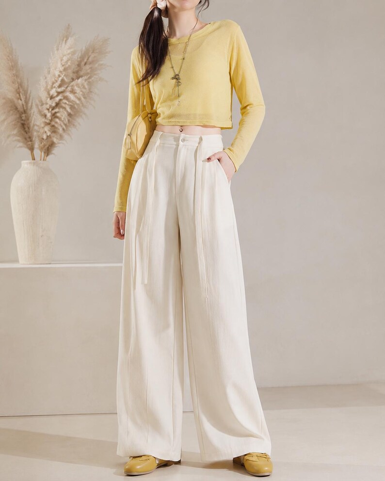 Puede incluir: Un conjunto femenino con un top corto amarillo de manga larga y pantalones de pierna ancha color crema. El conjunto se complementa con un peque&ntilde;o bolso amarillo y zapatos planos amarillos. Un jarr&oacute;n con plantas altas y secas en el fondo.