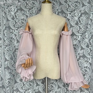 May include: Pale pink, off-the-shoulder sleeves with ruffled cuffs and a flowing design. The sleeves are displayed on a mannequin with wooden arms. The fabric appears sheer and lightweight, ideal for formal wear or bridal attire.