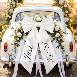 Peut inclure: Une voiture blanche vintage décorée pour un mariage. Un grand nœud en satin blanc et des compositions florales ornent l'arrière. Des rubans blancs et des bannières avec "Just Married" en écriture élégante complètent le look festif.