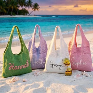 May include: Four knitted beach tote bags in green, lilac, cream, and pink, each with a name embroidered on the front. The bags are on a sandy beach with seashells and sunglasses. The background shows the sea and palm trees.