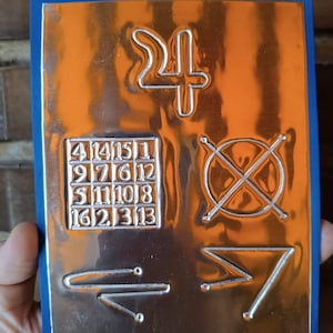 Puede incluir: Placa metálica rectangular con borde azul y centro naranja. La placa presenta símbolos en relieve, incluido el número 24, un cuadrado con números y otras formas geométricas. La placa está sujeta por una mano.
