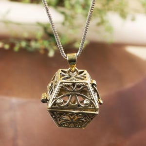 May include: A gold-colored filigree pendant necklace. The pendant is a geometric shape with intricate openwork designs. The necklace chain is silver-colored. The pendant has a hinged opening. The background is blurred, with hints of green and brown.