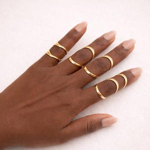 Puede incluir: Una mano que muestra varios anillos dorados. Los anillos tienen diseños variados, desde bandas simples hasta formas curvas. Los anillos se usan en varios dedos, creando un aspecto moderno y elegante. Los anillos son de oro.