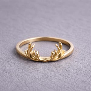 May include: A gold-colored ring featuring a detailed antler design. The ring has a smooth, polished band and the antlers are intricately crafted. The ring is set against a neutral gray background, highlighting its elegant design.