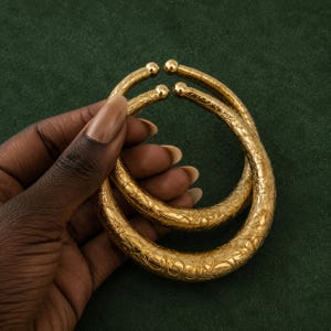 May include: Two gold-toned metal bracelets with an open-ended design and rounded ball terminals. The surface of each bracelet features an intricate, engraved floral pattern that covers the entire exterior of the thick, curved metal bands.