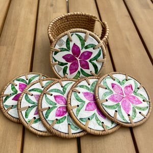 Könnte beinhalten: Set aus sechs dekorativen Untersetzern mit floralem Design. Jeder Untersetzer hat einen weißen Hintergrund mit einer rosa Blume und grünen Blättern, umrahmt von einem gewebten Rattanrand. Ein passender Rattan-Korb hält die Untersetzer.