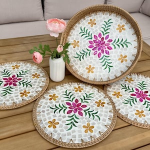 Könnte beinhalten: Ein Set aus vier dekorativen Rattan-Platzsets und einem passenden Serviertablett. Jedes Stück hat ein weißes Mosaikfliesen-Design mit einer zentralen rosa Blume, grünen Blättern und gelben Blüten. Die Ränder sind mit gewebtem Rattan verziert.