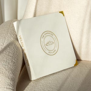 May include: A white wedding guest book with gold lettering and accents. The cover features a gold circular design with two swans and the text "THE JOHNSONS" and "JULY 6, 2020". The spine reads "HELEN & WALTER" in gold.