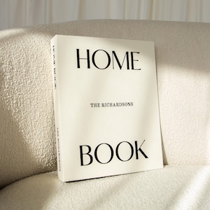 Pode incluir: Um livro de capa dura intitulado "HOME BOOK" com o texto "THE RICHARDSONS" em um design minimalista. O livro é branco sujo com letras pretas e está sobre uma superfície texturizada creme.