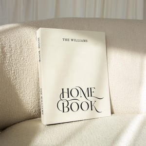 Op de afbeelding: Een crèmekleurig hardcover boek met de titel THE WILLIAMS HOME BOOK. De voorkant bevat de tekst THE WILLIAMS bovenaan en HOME BOOK in grote, gestileerde zwarte letters. De rug toont ook de tekst HOME BOOK en THE WILLIAMS.