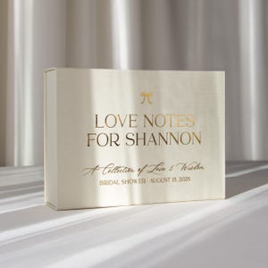 Puede incluir: Una caja de recuerdos rectangular de color blanco roto con texto en lámina dorada que dice LOVE NOTES FOR SHANNON, A Collection of Love & Wisdom, y BRIDAL SHOWER AUGUST 15, 2026, con un pequeño icono de lazo dorado sobre el título.