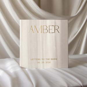 Puede incluir: Un libro cuadrado de color blanco roto con letras de lámina dorada en la portada. El nombre AMBER aparece en letras mayúsculas grandes y elegantes, con el texto LETTERS TO THE BRIDE y la fecha 06.28.2026 en una fuente dorada más pequeña.