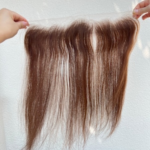 以下が含まれることがあります： 長いストレートの髪の毛が付いた茶色のヘアピース。ヘアピースは白い背景に対して両手で持たれています。髪の毛は赤茶色に見えます。