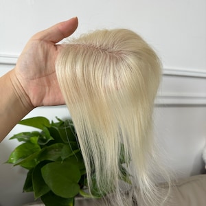 May include: A hair topper featuring straight, platinum blonde strands attached to a fine, skin-toned lace base. The hair is parted in the center, showcasing a realistic scalp appearance where the individual strands are knotted into the mesh material.