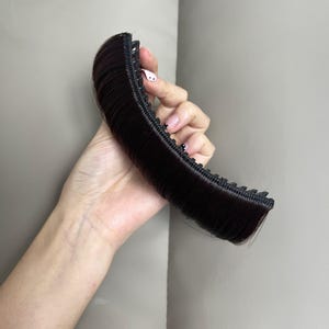 Pode incluir: Uma extensão de cabelo castanho escuro com uma base de pente de plástico preto curvo com dentes integrados. O cabelo sintético é estilizado numa franja lisa e arredondada, fixada de forma segura ao pente para uma aplicação fácil.