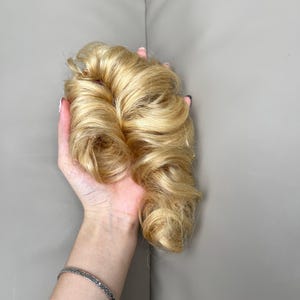 May include: A hair extension piece featuring voluminous, wavy, golden-blonde synthetic hair strands. The hair is styled in loose, soft curls that create a textured, layered appearance with varying shades of light and honey blonde throughout the piece.