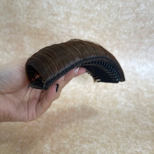 Pode incluir: Uma peça de extensão de cabelo castanho com uma base curva tipo pente com múltiplos dentes de plástico preto. O cabelo sintético é liso, reto e sobreposto à estrutura do pente, concebido para adicionar volume ou franja a um penteado.