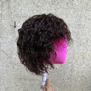 May include: A dark brown wig with tight, defined curls and a layered cut. The hair is styled on a bright pink mannequin head, showcasing a voluminous texture and a natural-looking wave pattern throughout the entire length of the dark brown strands.