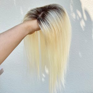 Puede incluir: Una pieza de cabello rubio con raíces oscuras se sostiene contra un fondo blanco. El cabello es largo y liso, con un degradado de color de aspecto natural. La pieza de cabello parece estar hecha de cabello humano.