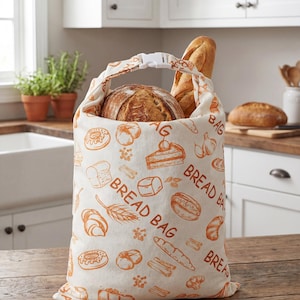 May include: A cream-colored fabric bread bag featuring an orange printed pattern of various baked goods and the text BREAD BAG. The bag has a white plastic buckle closure at the top and contains a round loaf of bread and a long baguette inside.