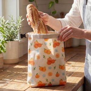 May include: A beige bread bag with an orange and floral pattern. A fresh baguette is being placed inside the bag. The bag has a black buckle closure and is sitting on a wooden surface.