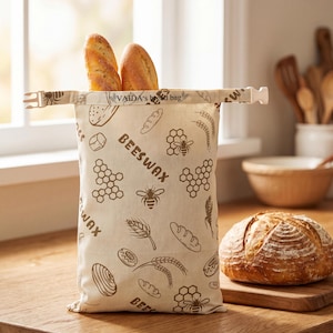 May include: A beige fabric bread bag with a brown print featuring bees, honeycombs, wheat stalks, and bread loaves. The text "VAIDA's bread bag" and "BEESWAX" are printed on the front. The bag has a plastic buckle closure at the top opening.