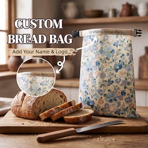 May include: A rectangular bread bag featuring a cream background with a blue and pink floral pattern. The bag has a roll-top closure with a black plastic buckle and the text VAIDA'S bread bag printed on the top edge. A loaf of bread is nearby.