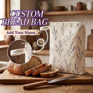 May include: A cream-coloured fabric bread bag featuring a lavender floral pattern and a zip closure. A small fabric tag on the side displays the name VAIDA. The bag is positioned next to a sliced loaf of bread on a wooden cutting board.