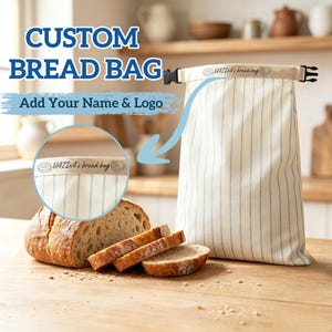 May include: A cream-colored bread bag with thin vertical blue stripes, featuring a roll-top closure with a black plastic buckle. The top band is printed with the text "VAIDA's bread bag" flanked by two small donut icons.