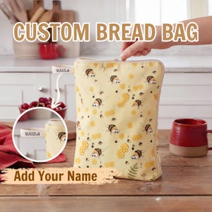 May include: A rectangular fabric bread bag with a light yellow background featuring a repeating pattern of cartoon bees and honeycombs. The bag includes a small side tab with the name VAIDA printed on it and a zipper closure along the top edge.
