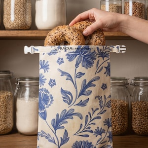 Op de afbeelding: Een rechthoekige broodzak met een crèmekleurige achtergrond en een blauw bloemenpatroon. De sluiting aan de bovenkant heeft een witte plastic gesp en de tekst VAIDA's bread bag in het zwart gedrukt. Twee meergranen bagels worden erin geplaatst.