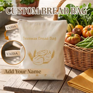 Op de afbeelding: Een crèmekleurige stoffen broodzak met ritssluiting en een zijlipje met het label VAIDA. De voorzijde bevat de tekst Beeswax Bread Bag, een afbeelding van brood en tarwe, en vier kleine iconen. De zak biedt ruimte voor een eigen naam.