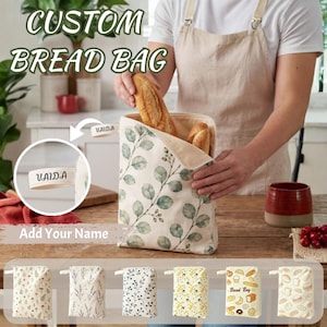 May include: A fabric bread bag featuring a green eucalyptus leaf pattern and a small side tab with the text VAIDA. The bag is open at the top, revealing two loaves of bread inside. A bottom row displays six additional bread bag design options.