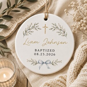 May include: A round white ceramic ornament featuring a gold cross with a heart center, green leaf illustrations, and the text Liam Johnson, BAPTIZED, and 08.23.2026. A light blue bow is printed at the bottom, and a jute twine loop is attached at the top.