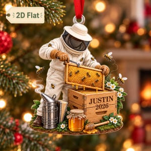 May include: A flat, decorative ornament featuring a beekeeper in a white protective suit holding a honeycomb frame over a wooden crate labeled JUSTIN 2026. The scene includes a metal smoker, a jar of honey, and several bees flying around.