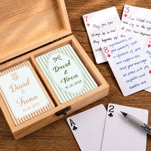 May include: A light wooden box containing two decks of personalised playing cards. One deck features a white and gold striped design with the text David & Fiona 10.30.2026. The other has green stripes and an oval frame with the same text.
