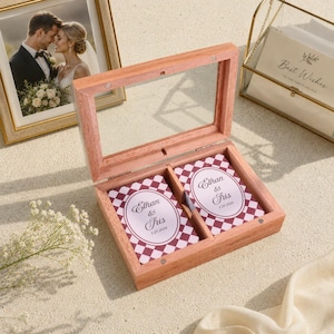 May include: A light-toned wooden box with a hinged glass lid containing two decks of playing cards. Each deck features a maroon and white checkered pattern with a central oval label printed with the text Ethan & Tris and the date 7.29.2026.