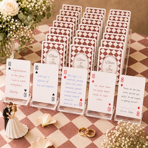 May include: A collection of custom playing cards displayed in clear acrylic stands on a checkered surface. Each card features a red and white diamond pattern on the back with the text Ethan & Iris, and handwritten messages on the front faces.