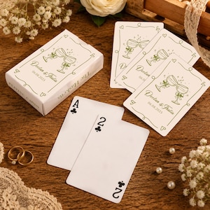 May include: A deck of white playing cards in a box, featuring a green line-art illustration of two clinking champagne glasses with bows. The text reads Declan & Thalia 06.08.2025 and The Hasketts. Two cards show the Ace and Two of clubs.