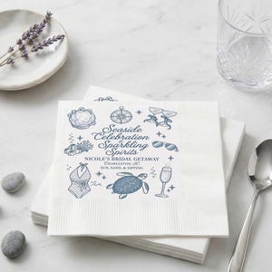 Pode incluir: Uma pilha de guardanapos de papel branco com um design azul impresso de uma bússola, tartaruga marinha, fato de banho, óculos de sol e flutes de champanhe. O texto diz Seaside Celebration & Sparkling Spirits, Nicole's Bridal Getaway, Charleston, SC, Sun, Sand, & Sipping.