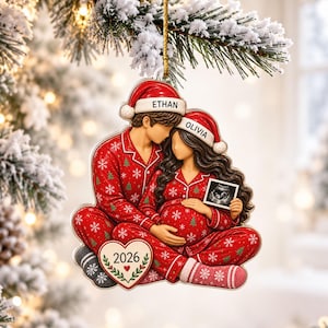 May include: A Christmas tree ornament featuring a couple in matching red snowflake pajamas and Santa hats labeled Ethan and Olivia. The figure holds an ultrasound image, and a heart-shaped plaque at the base displays the year 2026.