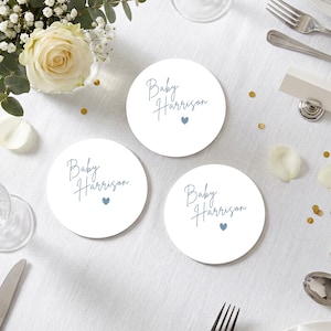 Può includere: Tre sottobicchieri circolari bianchi con il testo in corsivo blu Baby Harrison e una piccola icona a forma di cuore blu. I sottobicchieri sono disposti su una superficie bianca insieme a una rosa bianca, una forchetta, un cucchiaio, un coltello e coriandoli dorati.