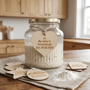 May include: A glass jar with a gold lid and a wooden heart tag that reads "Hi My name is: Fernandough Est. 04.08.2026." Several wooden tags and a pile of flour are on a linen cloth. The jar contains a bubbly substance.