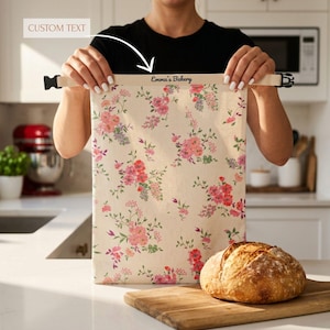 Könnte beinhalten: Ein rechteckiger Stoffbeutel mit cremefarbenem Hintergrund und einem rosa-grünen Blumenmuster. Die Oberkante hat ein beiges Band mit schwarzen Kunststoffschnallen an beiden Seiten und dem Text "Emma's Bakery" in schwarzer Schrift.