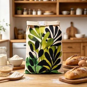 May include: A rectangular fabric storage bag featuring a cream-colored base with a bold, watercolor-style botanical print of large green and black leaves. The top of the bag is secured with a beige fabric strap and a white plastic buckle closure.