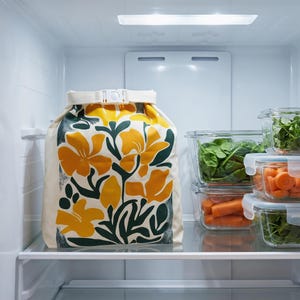 May include: A cream-colored roll-top storage bag featuring a bold floral pattern in mustard yellow and dark green. The bag is secured at the top with a white plastic buckle strap and is positioned on a refrigerator shelf next to glass food containers.
