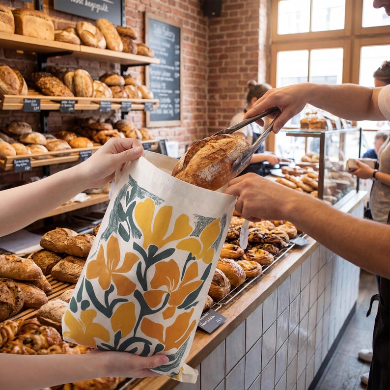 May include: A white fabric bag featuring a bold yellow and dark green floral pattern being filled with a crusty loaf of bread using metal tongs inside a bakery with wooden shelves stocked with various baked goods in the background.