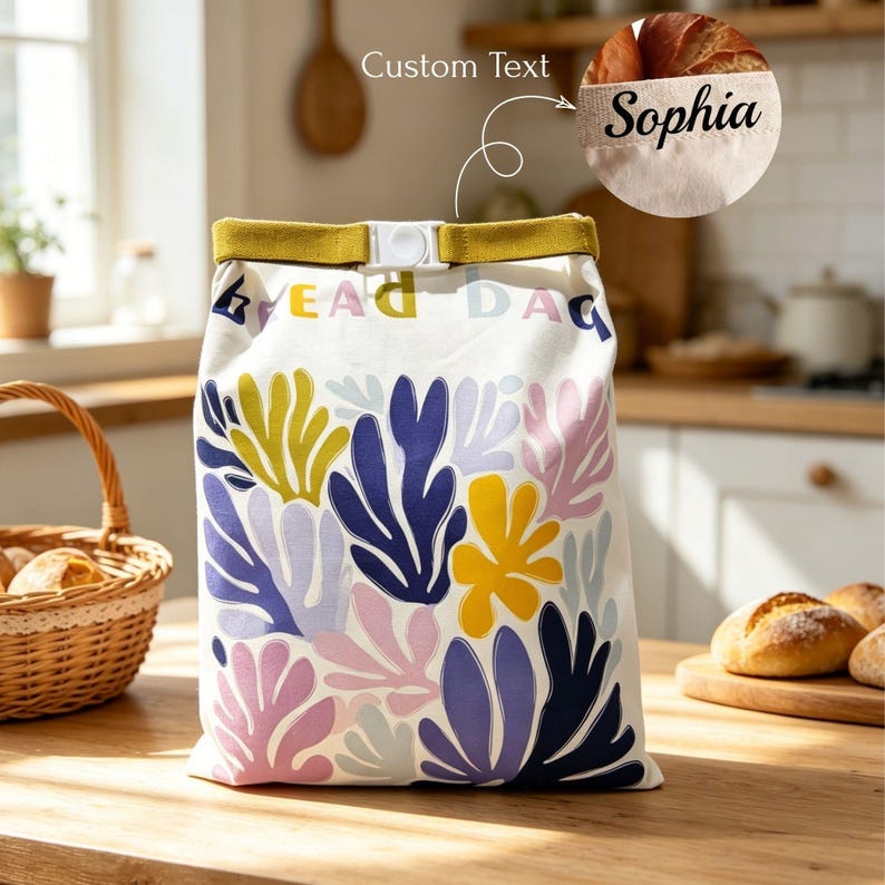 May include: A white fabric bread bag featuring a colorful pattern of abstract leaf shapes in blue, yellow, and pink. The bag has a mustard yellow roll-top closure with a white plastic buckle and the custom text Sophia printed on the top edge.