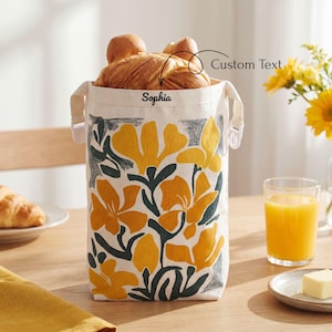 May include: A cylindrical canvas bread bag featuring a yellow and dark gray floral pattern. The bag has white side straps and the name Sophia printed in black text on the upper front. The bag is filled with golden brown baked bread rolls.