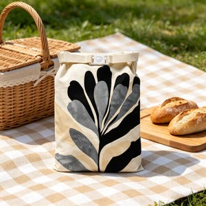 May include: A rectangular roll-top lunch bag featuring a cream-colored base with a bold, abstract botanical print in black and textured gray. The bag is secured at the top with a white plastic buckle closure and a matching cream-colored fabric strap.